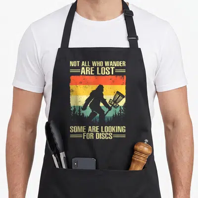 Player golf apron - Bigfoot silhouette holding a disc golf basket against a striped sunset with text — shown on a black cotton canvas apron against a white background