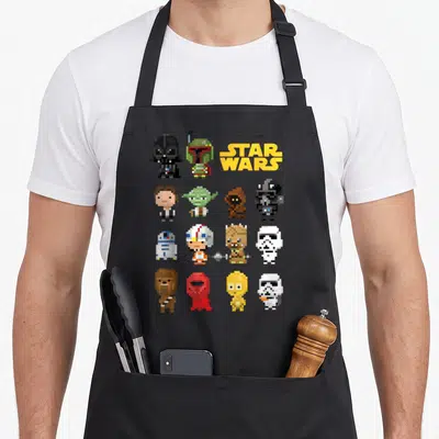 Heroe pixel apron - Pixel art grid of Star Wars characters with yellow logo text — shown on a black cotton canvas apron against a white background