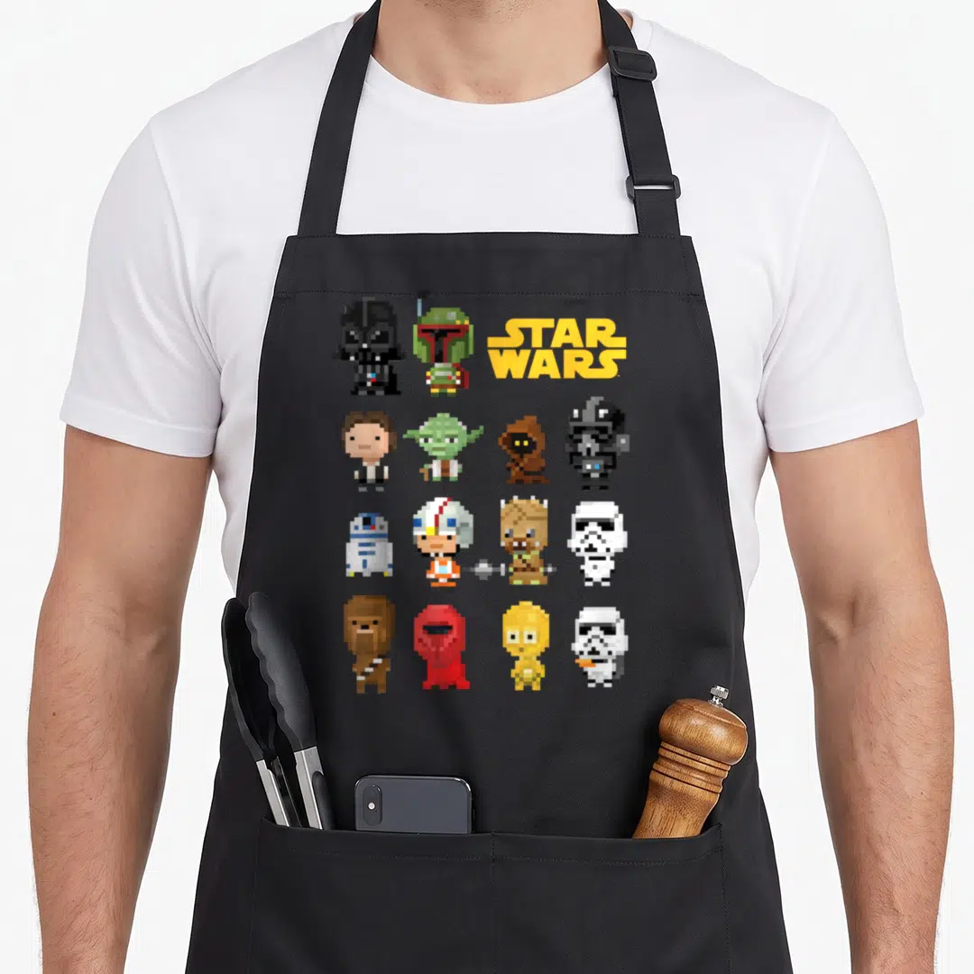 Heroe pixel apron - Pixel art grid of Star Wars characters with yellow logo text — shown on a black cotton canvas apron against a white background