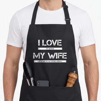 Pickleball wife apron - White stencil text reading I love it when my wife lets me play pickleball with horizontal bars — shown on a black cotton canvas apron against a white background