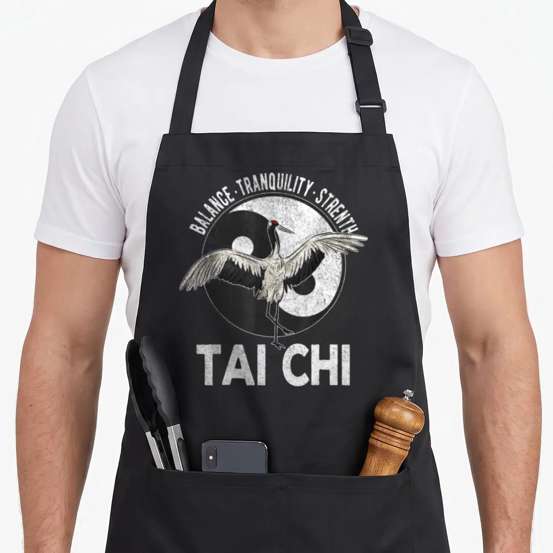 Chi crane apron - Distressed Tai Chi print with standing crane over yin yang symbol and arched text — shown on a black cotton canvas apron against a white background