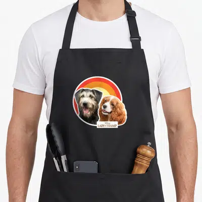 Live lady and the tramp apron - Two dogs beside red orange yellow rainbow with Disney text box — shown on a black cotton canvas apron against a white background