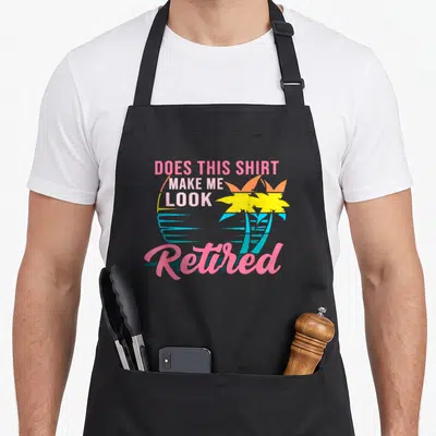 Cute doe retirement apron - Stacked Does This Shirt Make Me Look Retired text with two palm trees and sun in pink — shown on a black cotton canvas apron against a white background