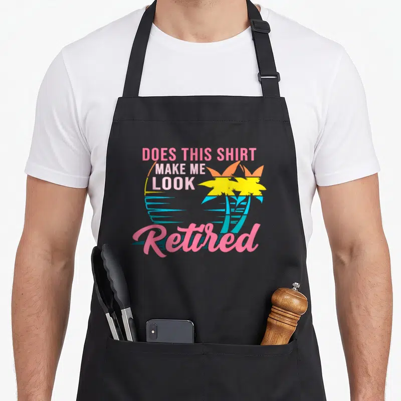 Cute doe retirement apron - Stacked Does This Shirt Make Me Look Retired text with two palm trees and sun in pink — shown on a black cotton canvas apron against a white background