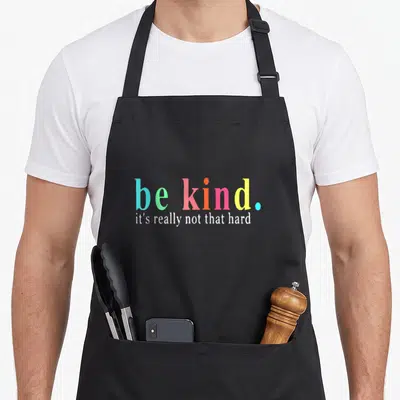 Kind hard apron - be kind it's really not that hard colorful stacked text design — shown on a black cotton canvas apron against a white background