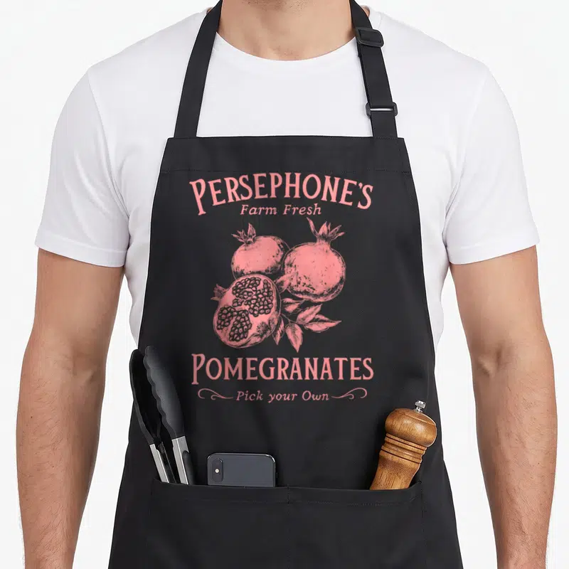 Dark pomegranate apron - Pink retro illustration of three pomegranates and leaves with arched text reading Persephone's — shown on a black cotton canvas apron against a white background