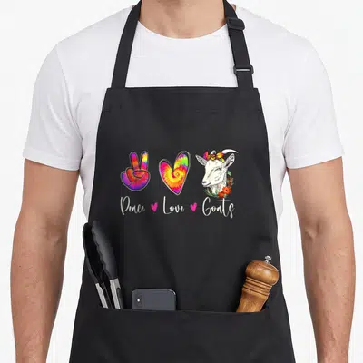 Peace goat apron - Tie-dye peace sign, heart, and goat head with floral wreath and pink hearts — shown on a black cotton canvas apron against a white background