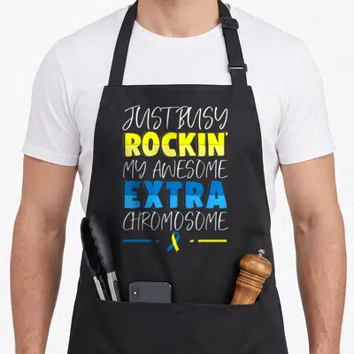 Funny down syndrome awareness apron - Stacked text reading Just Busy Rockin My Awesome Extra Chromosome above a blue — shown on a black cotton canvas apron against a white background