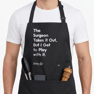 Scrub doctor apron - White stacked text reading The Surgeon Takes It Out But I Get to Play with it on a black background — shown on a black cotton canvas apron against a white background