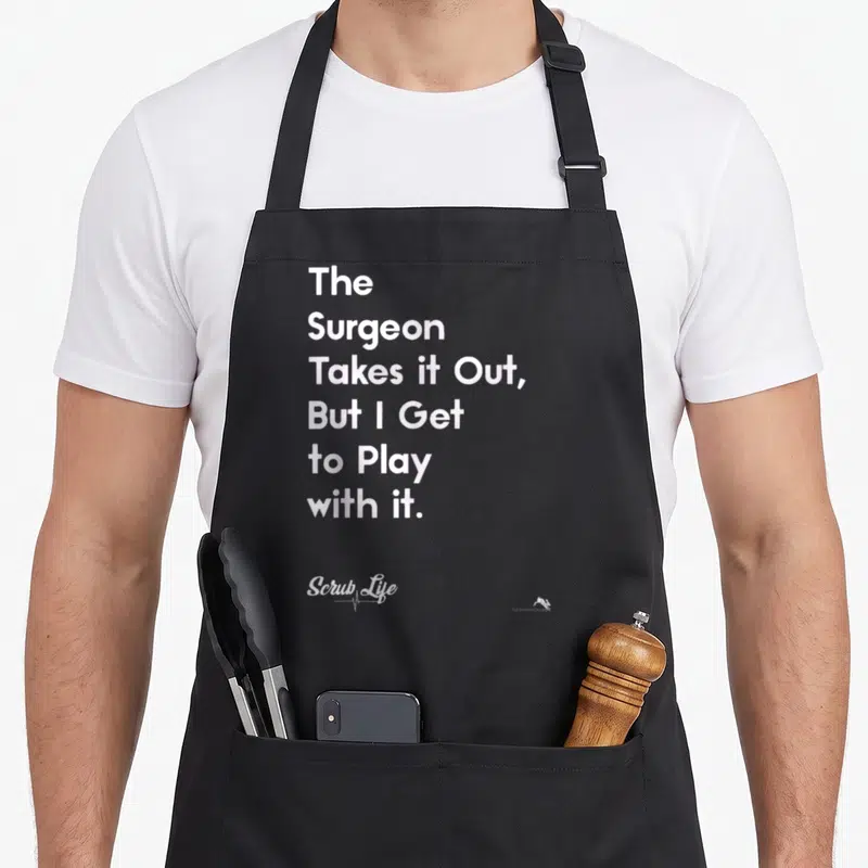 Scrub doctor apron - White stacked text reading The Surgeon Takes It Out But I Get to Play with it on a black background — shown on a black cotton canvas apron against a white background