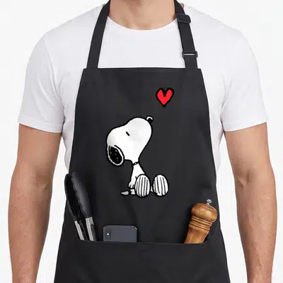 Sitting peanut apron - Cartoon white dog sitting looking up at a red heart shape — shown on a black cotton canvas apron against a white background