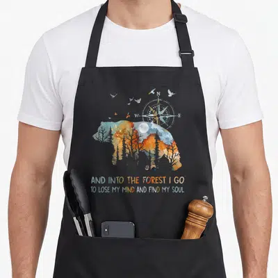 Forest camping apron - Watercolor bear silhouette containing forest scene with compass moon and birds above stacked — shown on a black cotton canvas apron against a white background