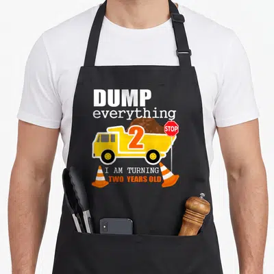2nd dump truck apron - Yellow dump truck with number 2, brown dirt, stop sign, and orange cones surrounding white — shown on a black cotton canvas apron against a white background