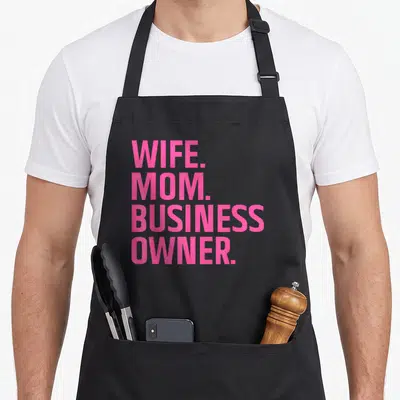 Wife small business aprons - Pink stacked text reading WIFE. MOM. BUSINESS OWNER. on a grey background — shown on a black cotton canvas apron against a white background