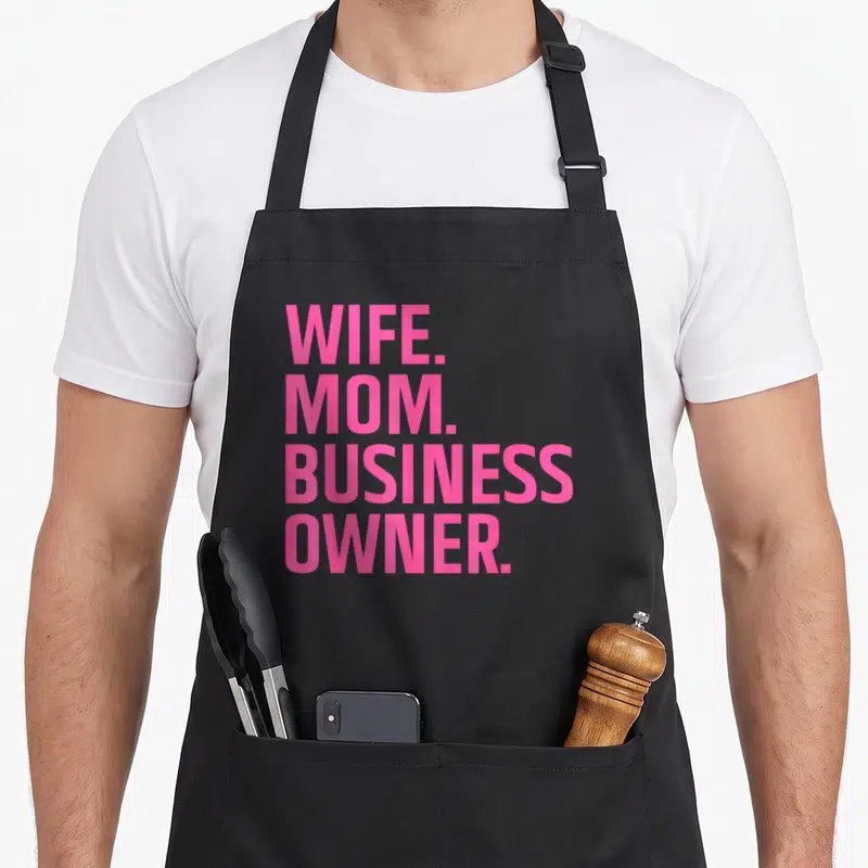 Wife small business aprons - Pink stacked text reading WIFE. MOM. BUSINESS OWNER. on a grey background — shown on a black cotton canvas apron against a white background