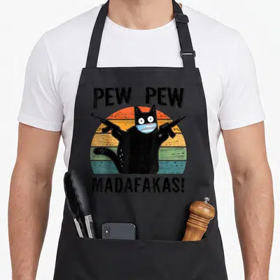 Pew cat apron - Black cat wearing blue mask holding two guns with PEW PEW MADAFAKAS text over striped sunset — shown on a black cotton canvas apron against a white background