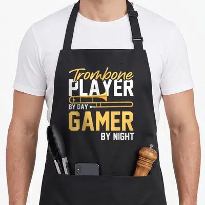 Player trombone apron - Yellow and white text reading Trombone Player By Day Gamer By Night with horizontal brass — shown on a black cotton canvas apron against a white background