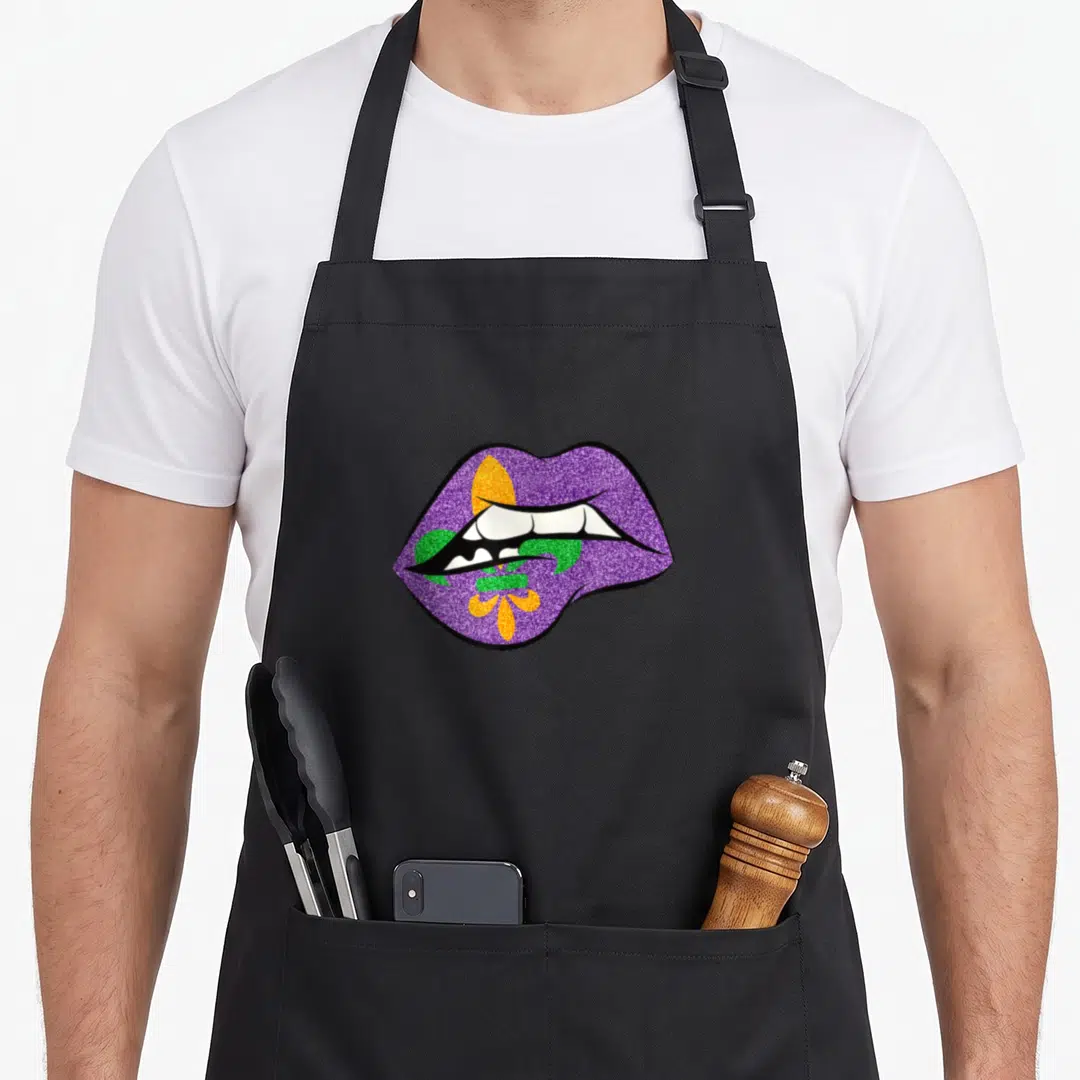 Lip fat chef apron - Glittery purple lips with overlapping gold and green fleur-de-lis symbol — shown on a black cotton canvas apron against a white background