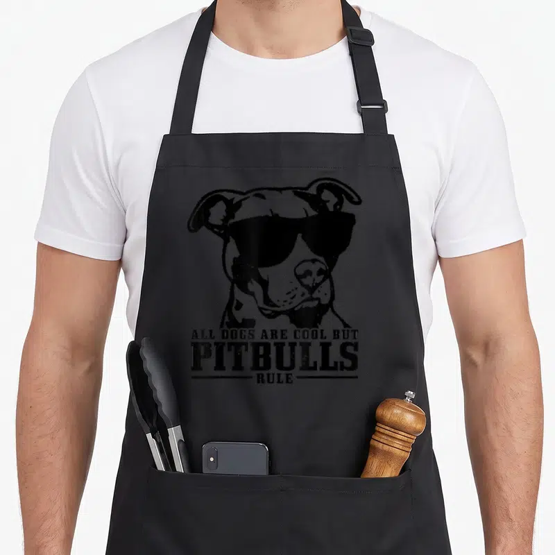 Pitbull dog rules apron - Black pitbull head wearing sunglasses above stacked distressed text reading All Dogs Are Cool — shown on a black cotton canvas apron against a white background