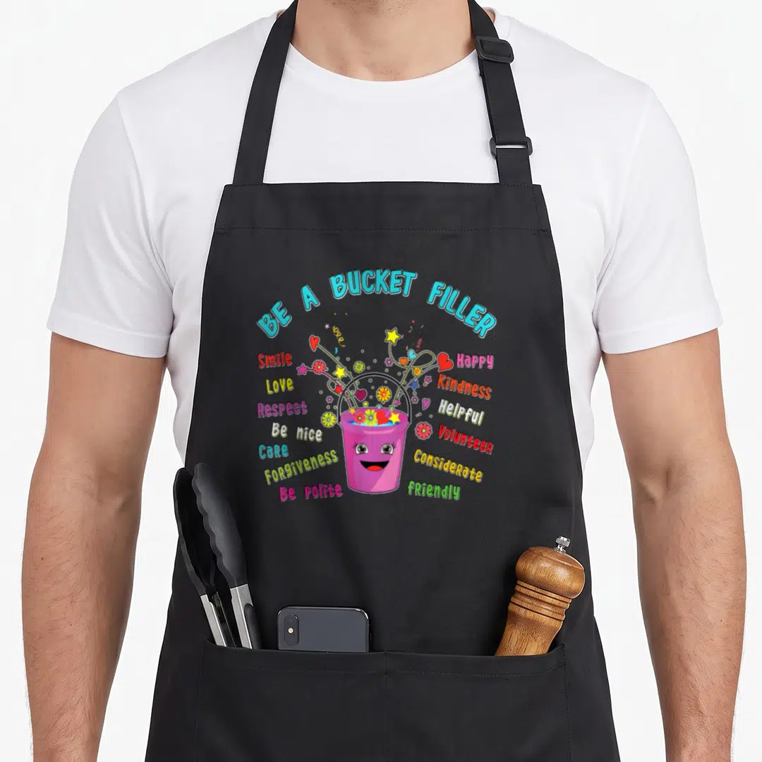 Guidance teacher apron - Pink smiling bucket with BE A BUCKET FILLER text and scattered virtues like kindness — shown on a black cotton canvas apron against a white background