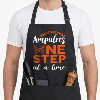 Loss ribbon apron - Orange and white text reading empowering amputees one step at a time with butterfly graphic — shown on a black cotton canvas apron against a white background