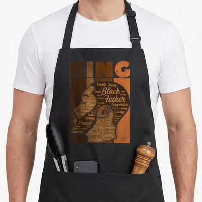 Father african american aprons - KING text above man profile silhouette filled with words like Noble, Swag, Proud — shown on a black cotton canvas apron against a white background