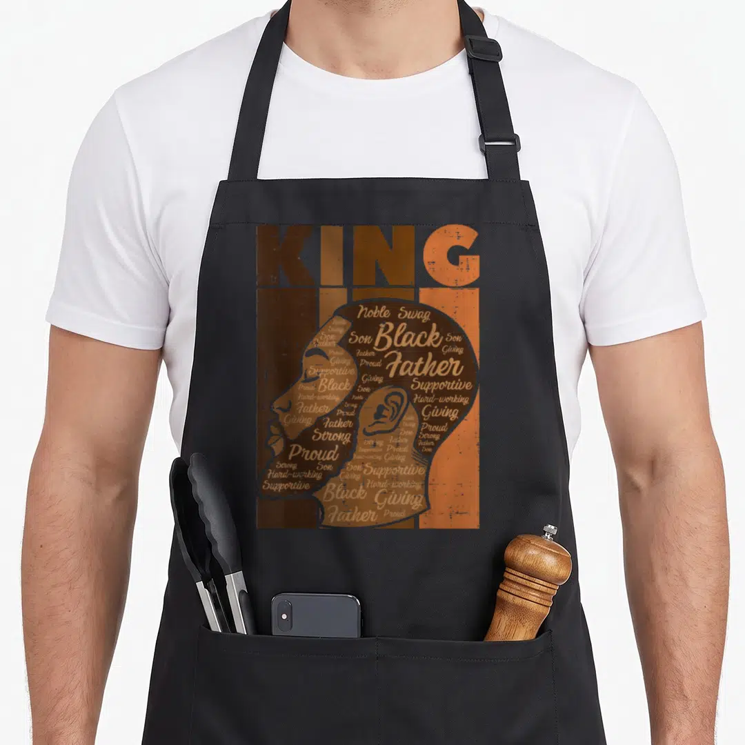 Father african american aprons - KING text above man profile silhouette filled with words like Noble, Swag, Proud — shown on a black cotton canvas apron against a white background