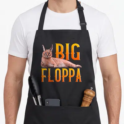 Floppa cat apron - Cartoon caracal lying between orange BIG and FLOPPA text in tan and yellow — shown on a black cotton canvas apron against a white background