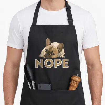 Nope lazy french bulldog apron - Tan cartoon french bulldog lying down above tan text reading NOPE with white outline — shown on a black cotton canvas apron against a white background