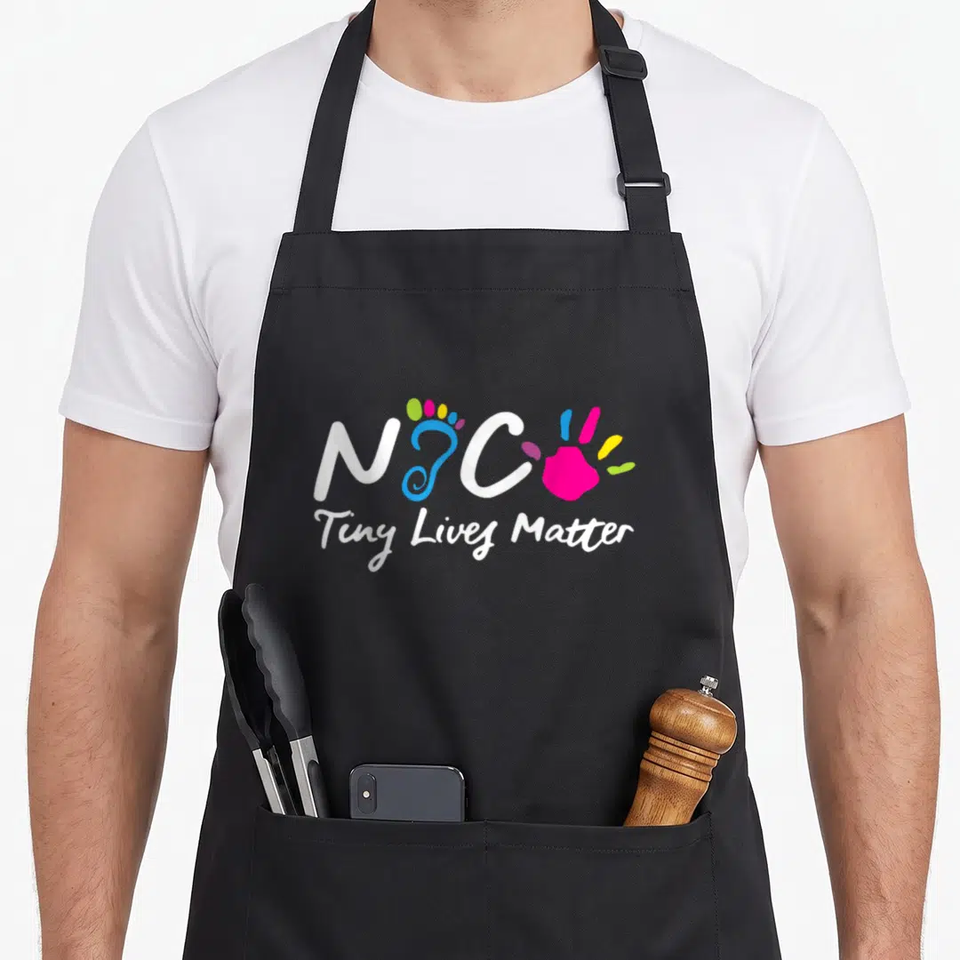 Taking tiny chef apron - NIC letters flanking colorful footprint and handprint with Tiny Lives Matter text below — shown on a black cotton canvas apron against a white background