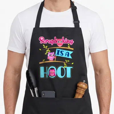 Scrapbooking owl apron - Three cartoon owls perched on branches with pink and blue Scrapbooking IS A HOOT text — shown on a black cotton canvas apron against a white background