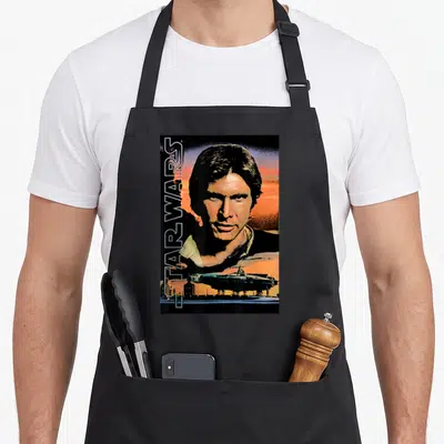 Poster falcon apron - Vertical Star Wars text, Han Solo portrait, Millennium Falcon, retro orange and blue art — shown on a black cotton canvas apron against a white background