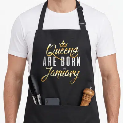 Cute january birthday apron - Gold and silver text reading Queens Are Born In January with a crown icon — shown on a black cotton canvas apron against a white background