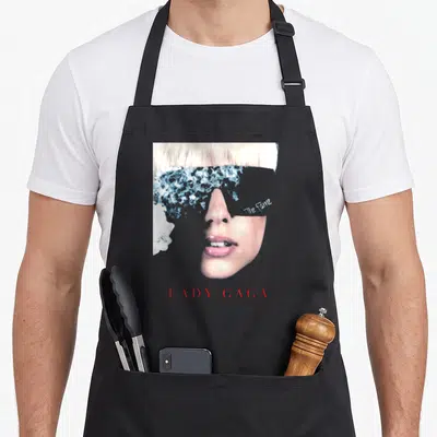 Gaga fame apron - Woman's face with blue pattern covering eyes, 'The Fame' on cheek, 'LADY GAGA' in red below — shown on a black cotton canvas apron against a white background