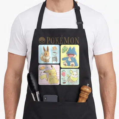 Munchlax eevee apron - POKÉMON text and four panels of Eevee, Munchlax, Pikachu, and Yamper with food items — shown on a black cotton canvas apron against a white background