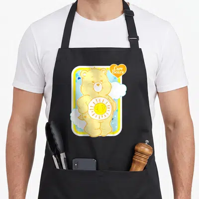 Funshine bear apron - Yellow bear with sun emblem and Care Bears text in orange heart — shown on a black cotton canvas apron against a white background