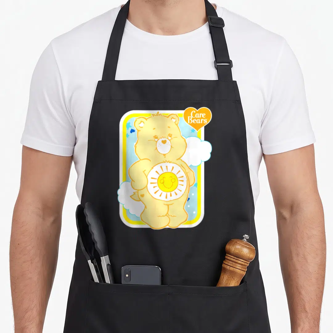Funshine bear apron - Yellow bear with sun emblem and Care Bears text in orange heart — shown on a black cotton canvas apron against a white background