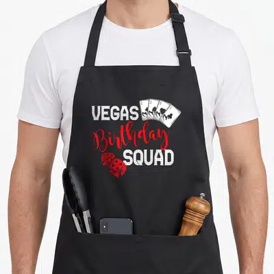 Funny vegas birthday apron - Stacked VEGAS Birthday SQUAD text with four playing cards and two dice in white and red — shown on a black cotton canvas apron against a white background