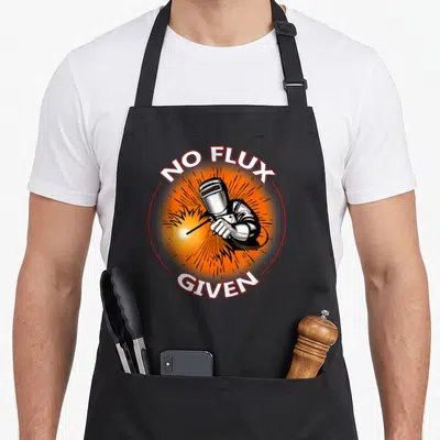 Funny flux welder dad apron - Cartoon welder in red circle with orange burst and text NO FLUX GIVEN — shown on a black cotton canvas apron against a white background