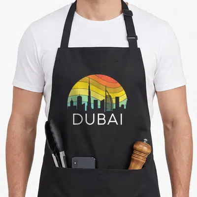 Dubai travel girl apron - Retro Dubai skyline with colorful sun stripes and white text — shown on a black cotton canvas apron against a white background