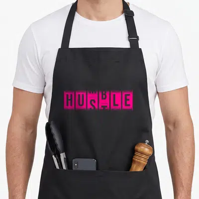 Humble hustle apron - Pink typography art showing HUSTLE in solid blocks with HUMBLE outlined over it — shown on a black cotton canvas apron against a white background