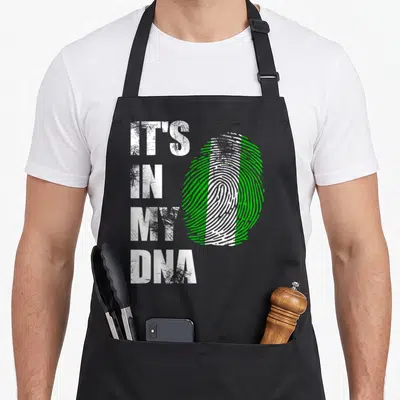Nigeria distressed apron - IT'S IN MY DNA text next to a white and green fingerprint shaped like a flag — shown on a black cotton canvas apron against a white background