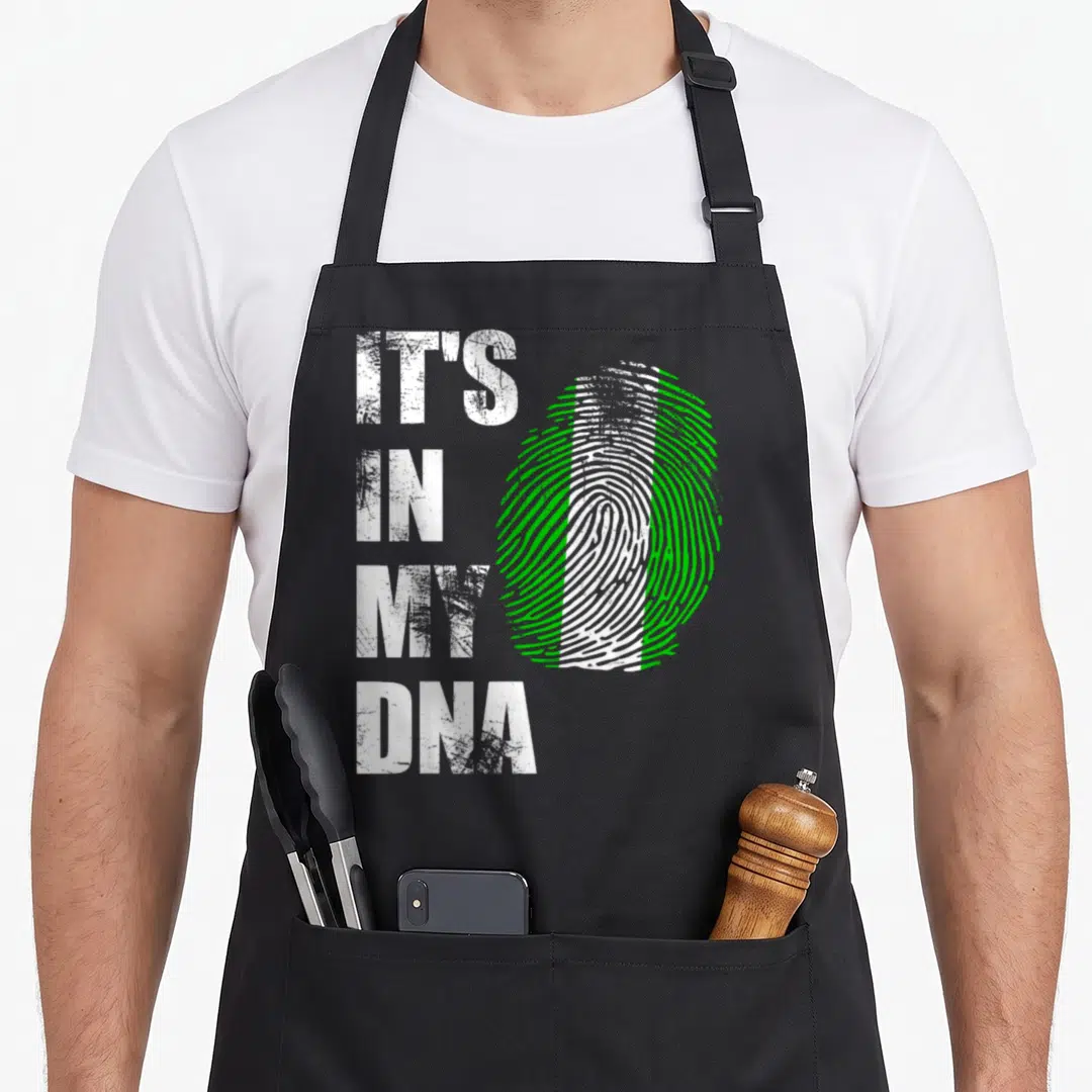 Nigeria distressed apron - IT'S IN MY DNA text next to a white and green fingerprint shaped like a flag — shown on a black cotton canvas apron against a white background
