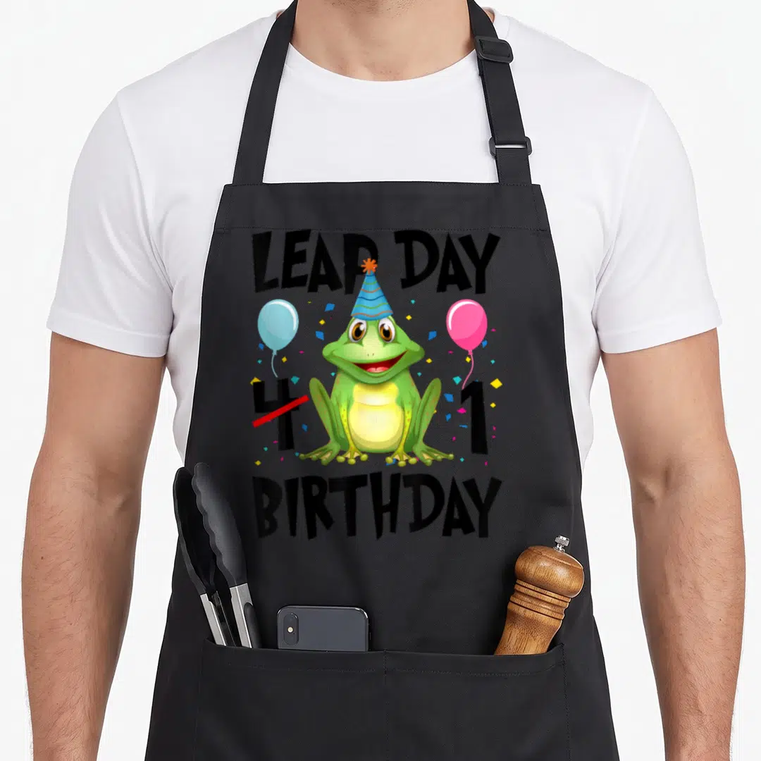 Leap 1st birthday apron - Green frog wearing blue party hat with balloons and text LEAP DAY 1 BIRTHDAY — shown on a black cotton canvas apron against a white background