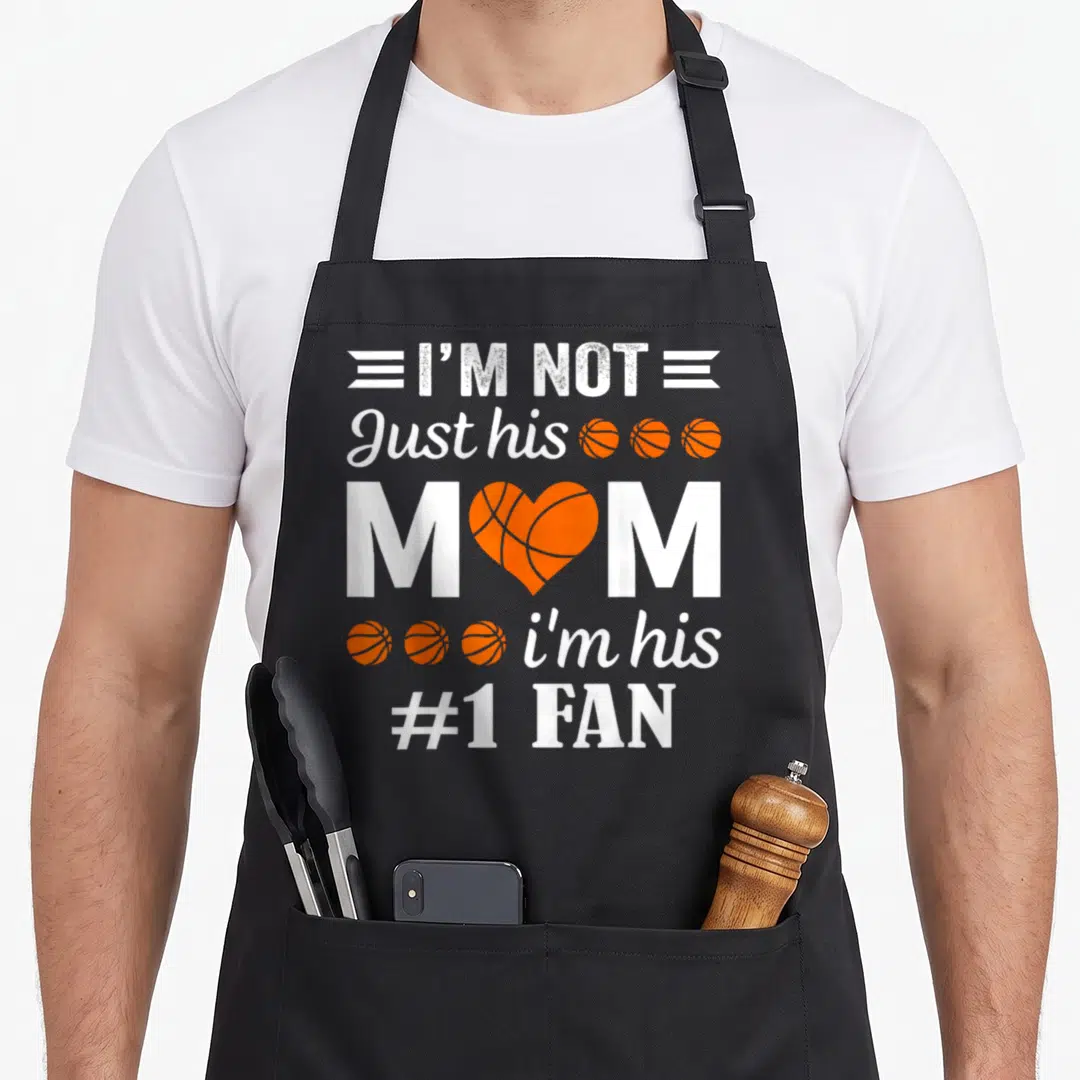Basketball fan quotes apron - Stacked white and orange text I'M NOT Just his MOM i'm his #1 FAN with heart basketball — shown on a black cotton canvas apron against a white background