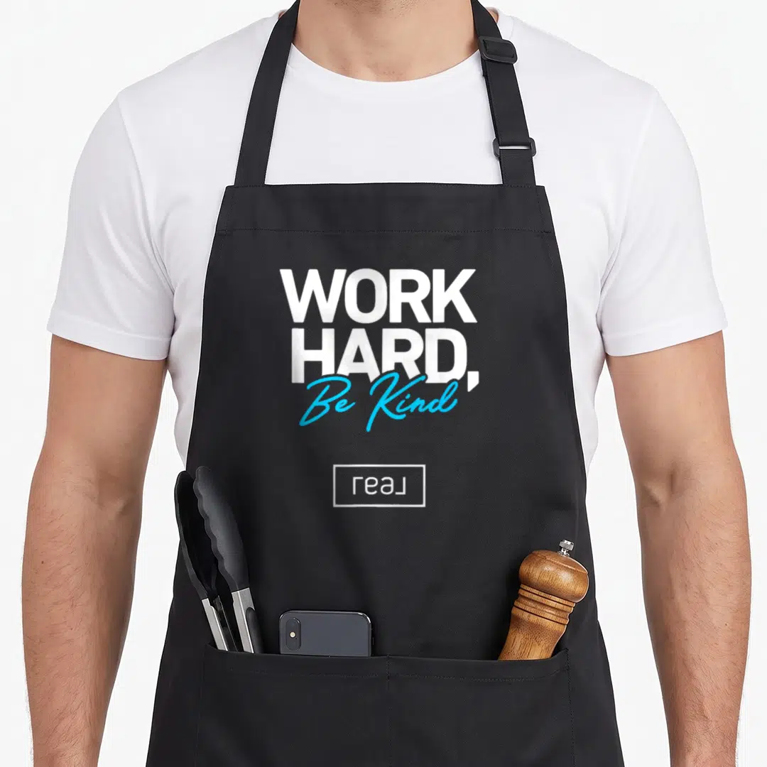 Broker work hard apron - White and blue Work Hard Be Kind typography with small boxed logo below — shown on a black cotton canvas apron against a white background