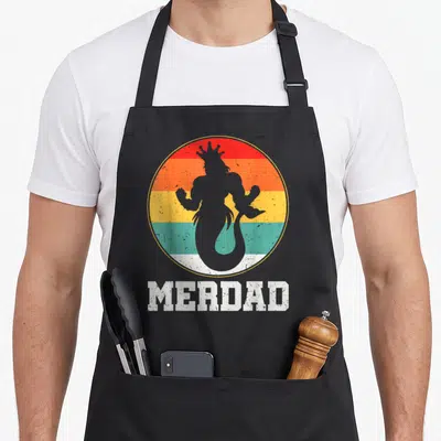 Merdad safety apron - Grey merman silhouette in red orange yellow teal striped circle with MERDAD text below — shown on a black cotton canvas apron against a white background