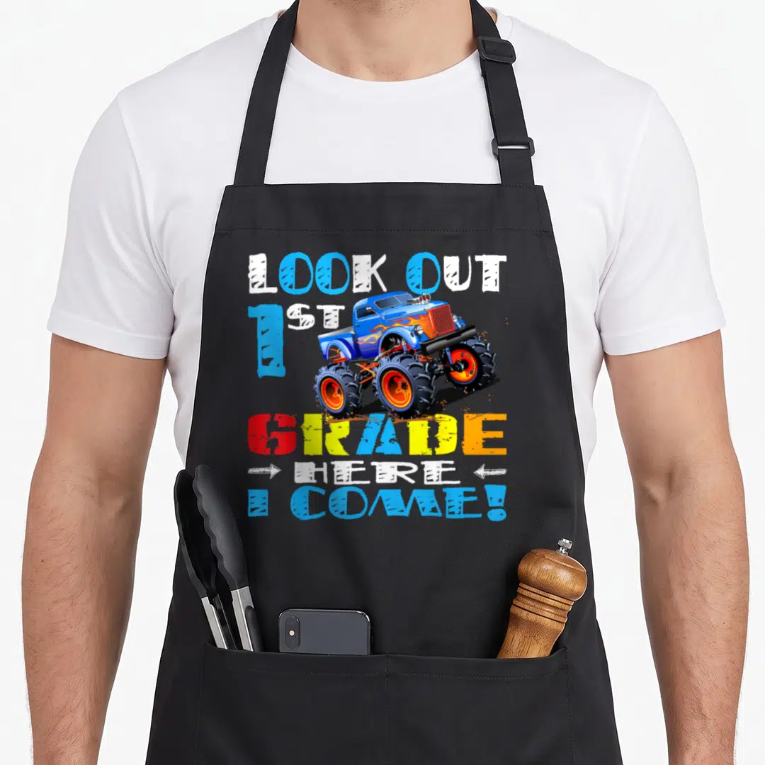 Come truck apron - Monster truck with LOOK OUT 1ST GRADE HERE I COME text in blue orange red yellow white cartoon style — shown on a black cotton canvas apron against a white background