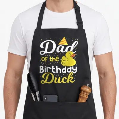 Birthday cotton duck apron - Dad of the Birthday Duck text with yellow rubber duck and striped party hat surrounded — shown on a black cotton canvas apron against a white background