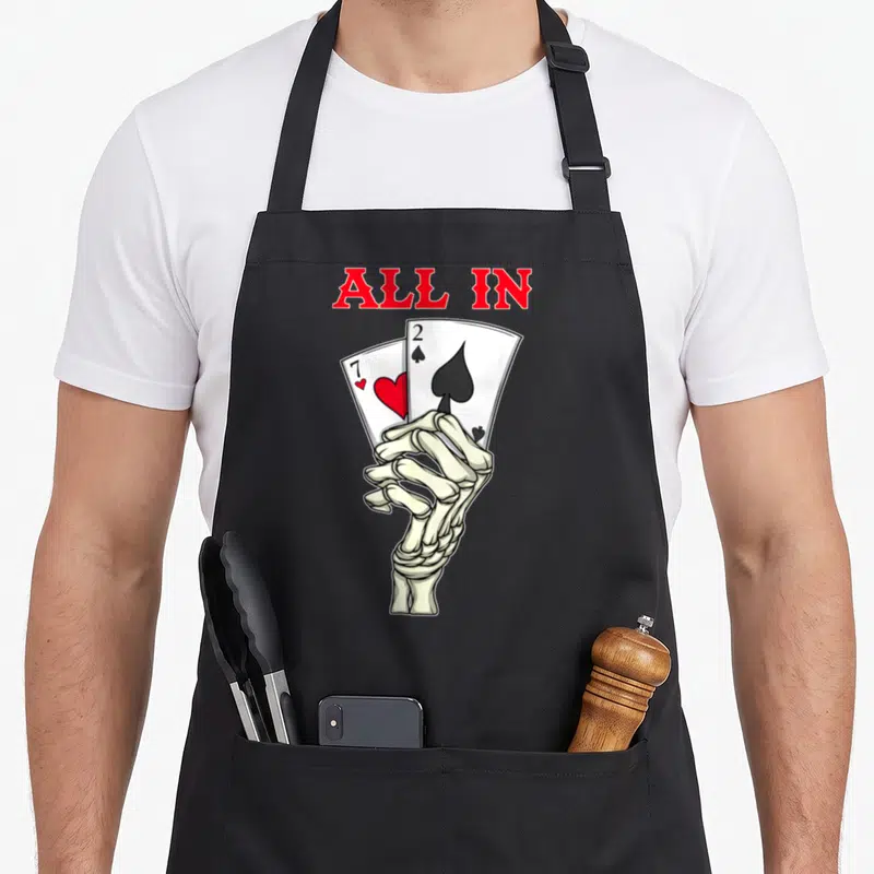 Poker skeleton apron - Skeleton hand holding 7 of hearts and 2 of spades below red ALL IN text — shown on a black cotton canvas apron against a white background